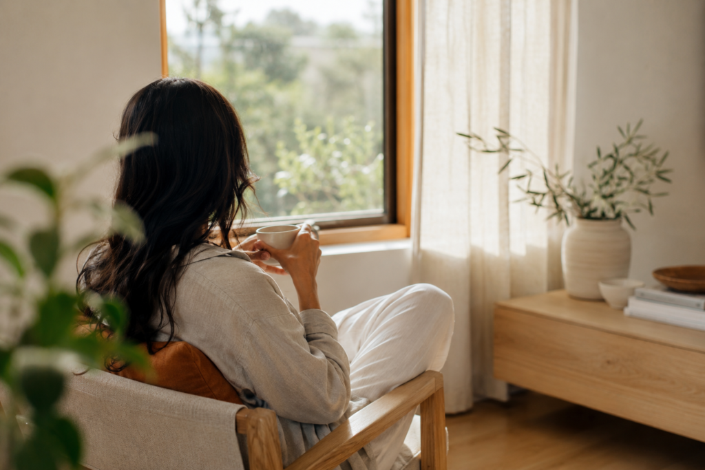 Woman sitting comfortably by a sunny window while holding a warm cup.